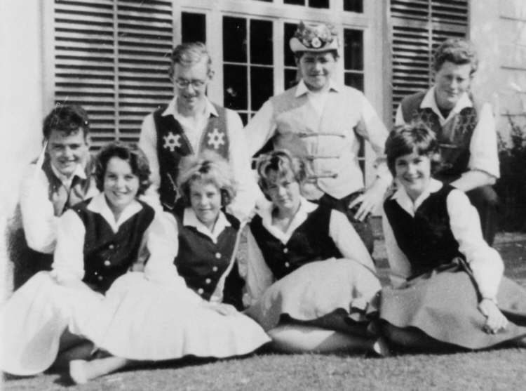 Young men in embroidered waistcoats and girls in dance skirts, sitting together on a lawn