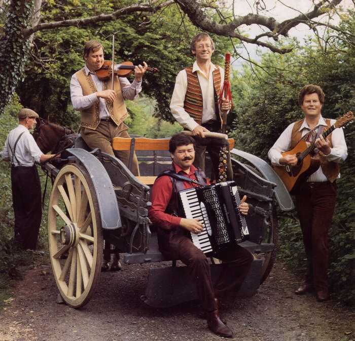 Musicians with a fiddle, an accordion and a guitar on and beside a horse drawn cart in a country lane
