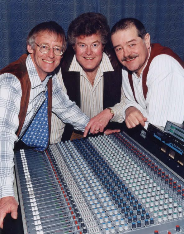 The three leaning over a large mixing desk in a recording studio