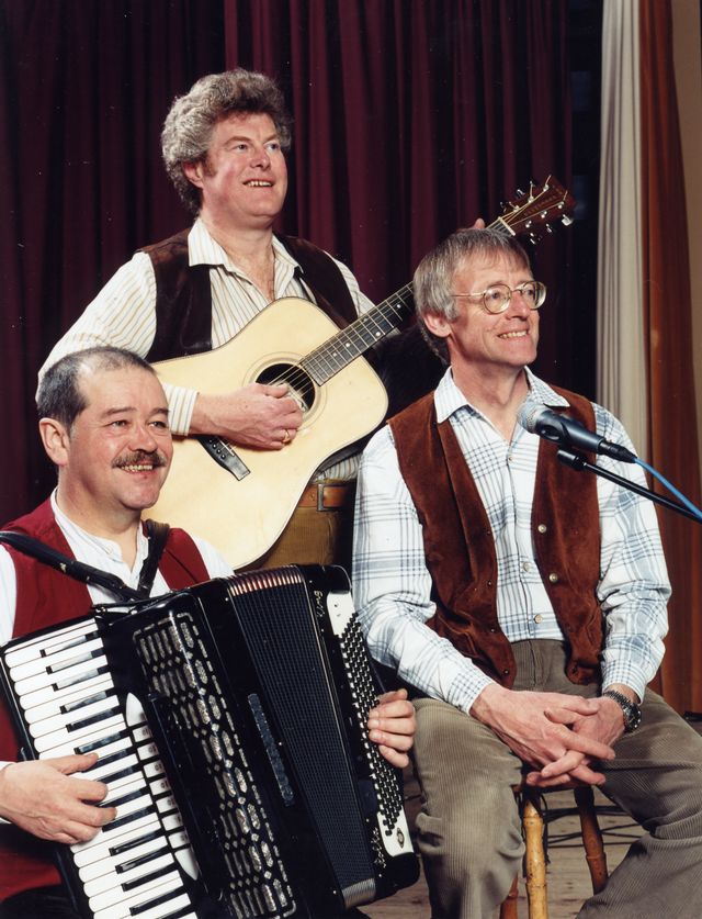 The three on stage with an accordion, a guitar and a microphone, in front of a red curtain