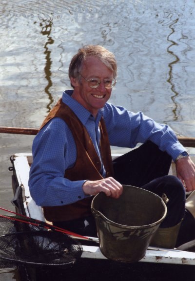 A man with glasses in a blue shirt and brown waistcoat, sitting in a boat beside a bucket