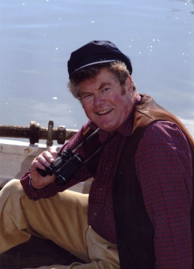 A man in a peaked cap holding binoculars, sitting in a boat on the water