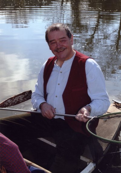 A man with a moustache in a red waistcoat, sitting in a boat and holding a landing net
