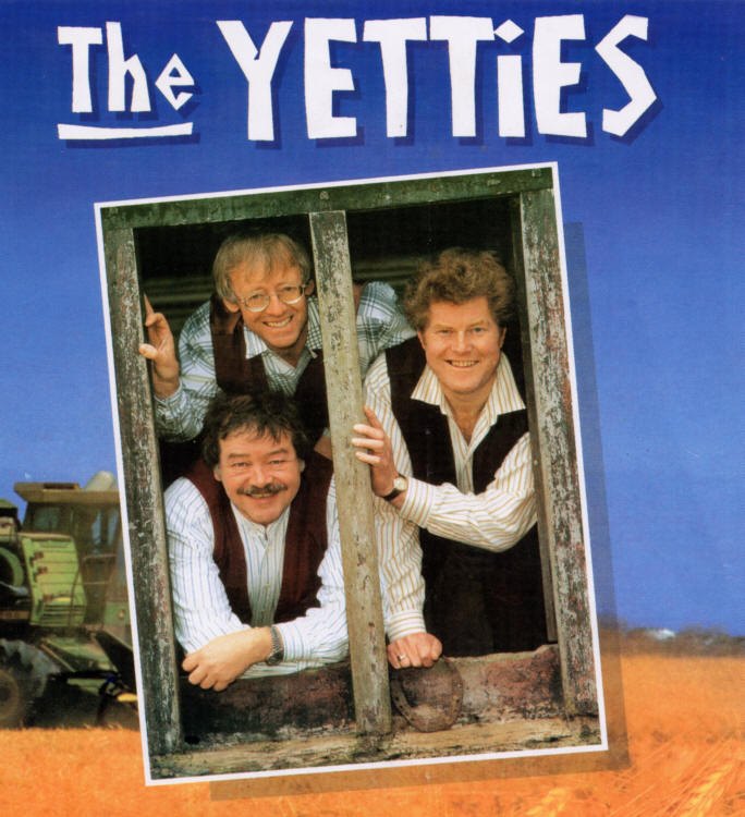 The three looking out through an old wooden window frame under The Yetties lettering, with a tractor behind and a cornfield below