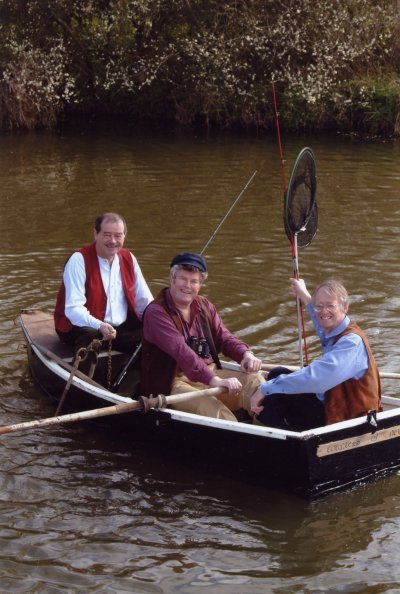 The three in a small rowing boat on a river, one holding up a landing net