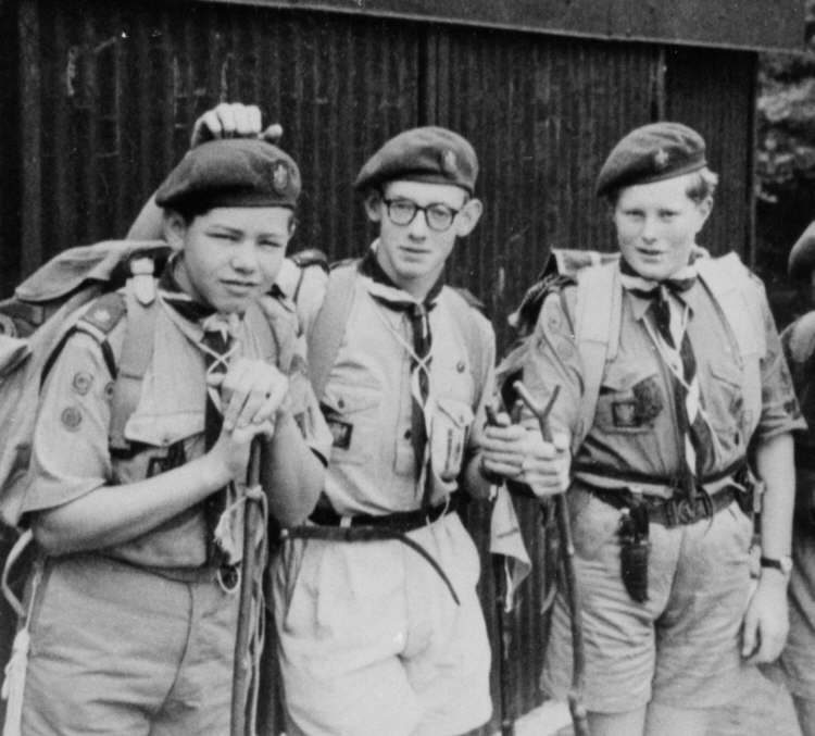 Three Scouts in berets with rucksacks and staffs, outside a corrugated iron hut